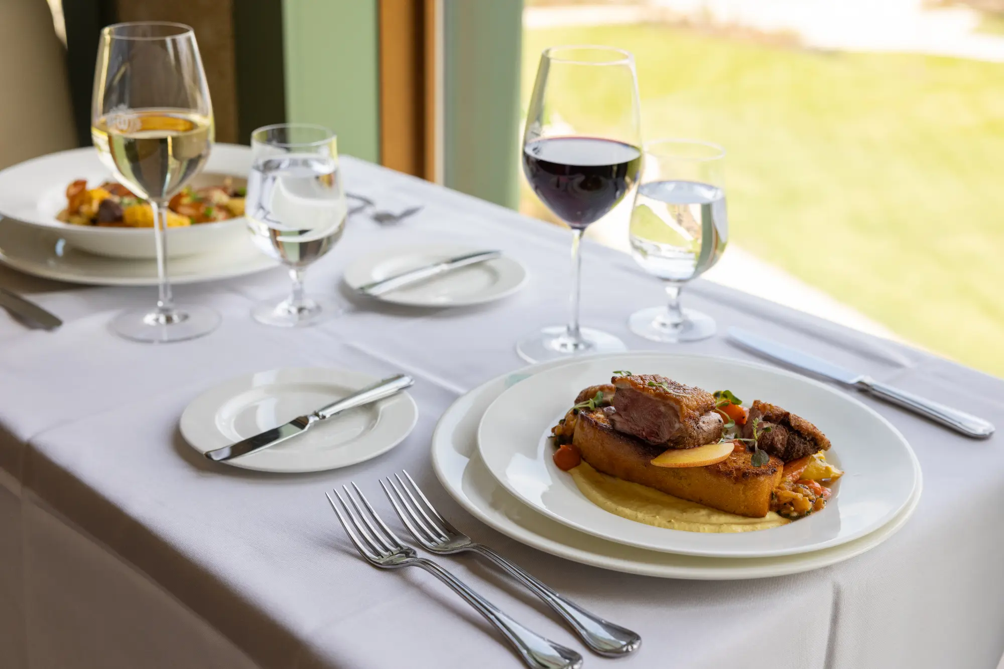 Fine dining table set with wine and plated entrées in a bright lakeside setting.